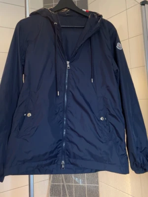 Mörkblå vindjacka från Moncler - Snygg mörkblå vindjacka från Moncler med huva och dragkedja framtill. Jackan har två fickor med tryckknappar, dragsko i huvan och en diskret Moncler-logga på ärmen. Materialet är tunt och lätt, perfekt för blåsiga dagar. Röd och vit detalj på huvan baktill.priset kan diskuteras vid snabb affär.| För fler frågor och funderingar hör av er.✨