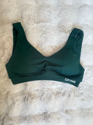 Mörkgrön sport-bh från DFYNE - Mörkgrön sport-bh från DFYNE med breda axelband och elastisk resår under bysten. Bh:n har en stilren design utan vaddering och logga framtill. Tillverkad i ett mjukt och stretchigt material som ger bra stöd.
