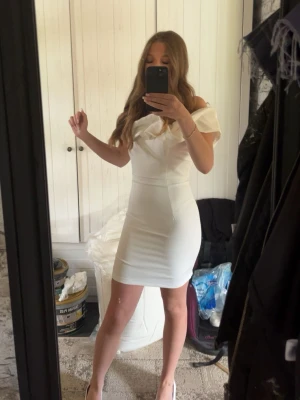 Vit offshoulder kort klänning - Snygg vit klänning med offshoulder-design och figurnära passform. Klänningen är kort och har en elegant v-ringning framtill samt öppen rygg. Ej genomskinlig då den har extra tyg under. Perfekt för dig som vill ha en stilren och modern look.