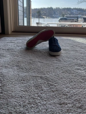 Marinblå sneakers med nitar - Säljer ett par marinblå sneakers från Christian Louboutin med klassiska röda sulor och coola nitar på sidorna. Skorna har en låg profil och är tillverkade i mocka med vita gummisulor. Perfekta för dig som vill sticka ut med en edgy och lyxig stil.
