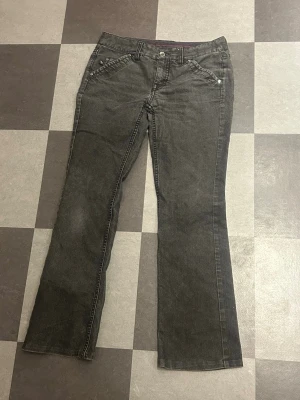 Mörkgrå bootcut jeans med detaljer - Snygga mörkgrå jeans med bootcut passform och coola silverfärgade nitar vid fickorna. Klassisk femficksdesign och knappstängning framtill. Perfekta för dig som vill ha en avslappnad men ändå stilren look.