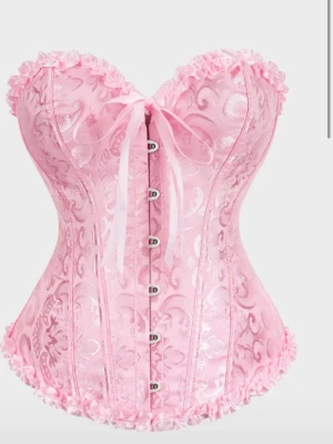 Rosa korsett med spets och rosett - Supersöt rosa korsett med glänsande mönster, spetsdetaljer och en dekorativ rosett framtill. Korsetten har hakknäppning fram och volangkant längs över- och nederkant.