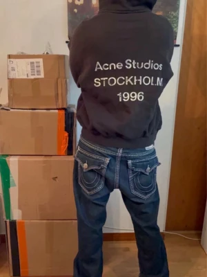 Svart hoodie Acne Studios Stockholm - Svart hoodie från Acne Studios med stor vit text på ryggen: 'Acne Studios STOCKHOLM 1996'. Klassisk passform med huva och ribbade muddar. Perfekt för dig som gillar streetwear och vill ha en stilren look med tydlig logga.
