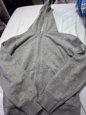 Grå zip-hoodie med lång huva - En stilren grå hoodie med dragkedja och extra lång huva. Tröjan har ribbade muddar vid ärmslut och nederkant samt fickor framtill. Perfekt för en avslappnad look. Materialet är mjukt och bekvämt, troligtvis bomullsblandning.