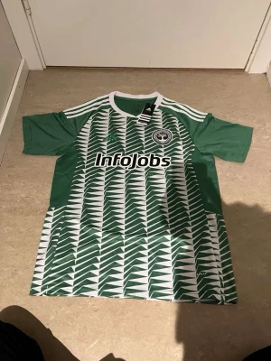 Grön Adidas fotbollströja InfoJobs - Säljer en grön och vit fotbollströja från Adidas med InfoJobs-logga på bröstet och ett mönster av vita pilar framtill. Tröjan har rund halsringning, korta ärmar och klubbmärke på vänster bröst. Perfekt för träning eller match!