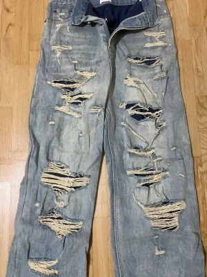 Ljusblå ripped jeans från Mijko - Säljer ett par ljusblå jeans från Miuko med rejält slitna och trasiga detaljer framtill. Jeansen har en avslappnad passform och är tillverkade i klassisk denim. Perfekta för en edgy streetstyle-look. 