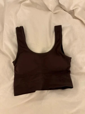 Mörkbrun ribbad sport-bh - Snygg mörkbrun sport-bh med breda axelband och ribbad nederkant. Bh:n har en enkel, stilren design och är tillverkad i ett stretchigt syntetmaterial. Perfekt för dig som vill ha en bekväm och smidig bh utan bygel.