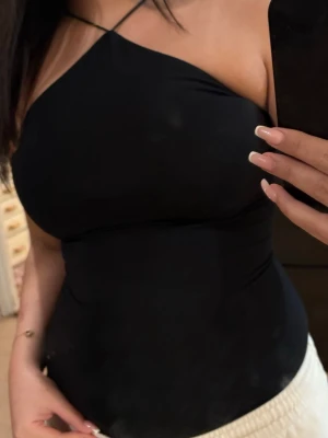 Svart oneshoulder topp med smala band - Snygg svart oneshoulder topp med tunna axelband och tight passform. Perfekt för dig som vill ha en stilren och trendig look. Toppen är ärmlös och har en enkel, clean design som passar till många olika outfits.