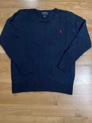 Mörkblå stickad tröja Polo Ralph Lauren - Mörkblå stickad tröja från Polo Ralph Lauren med klassiskt kabelstickat mönster och rund halsringning. Tröjan har ett rött broderat logotyp på bröstet och ribbade muddar vid ärmslut och nederkant. Perfekt för dig som gillar stilrena plagg.