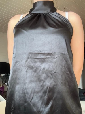 Svart halterneck topp i satin - Snygg svart halterneck topp i glansigt satinmaterial med hög krage och öppen rygg. Toppen knyts i nacken och har en loose passform som ger en cool och elegant look. Perfekt för att skapa en trendig outfit.