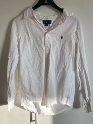 Vit skjorta från Ralph Lauren - Klassisk vit skjorta från Ralph Lauren med lång ärm och knappar framtill. Skjortan har den ikoniska broderade loggan på bröstet och är tillverkad i bomull. Perfekt till en clean och stilren look. Storlek xs men passar bra på s