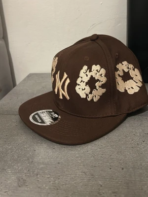 Brun New Era keps med brodyr - Snygg brun keps från New Era med platt skärm och broderade vita detaljer samt NY-logga framtill. Kepsen har justerbar passform och är tillverkad i bomullsmaterial. Perfekt för dig som vill sticka ut med en unik accessoar.