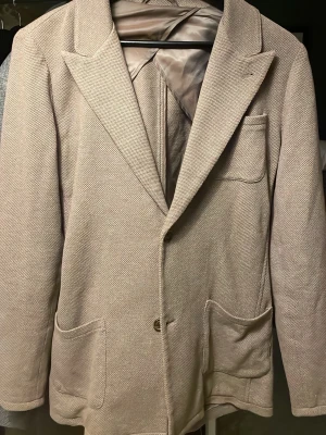 Beige kavaj från John Henric - Stilren beige kavaj från John Henric i 100% bomull. Klassisk modell med breda slag, tre utanpåfickor och en bröstficka. Perfekt för en dressad look med en modern touch. Fodrad insida för extra komfort.