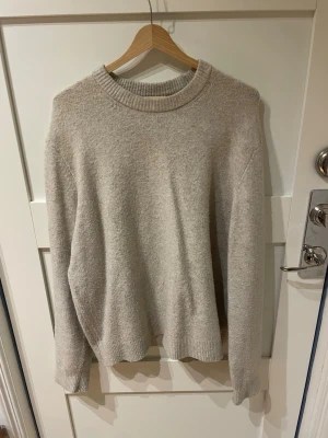 Beige stickad tröja i ullmix - Mysig beige stickad tröja med rund halsringning och ribbade muddar vid ärmslut och nederkant. Tröjan har en avslappnad passform och är perfekt för lager på lager. Storlek: M.