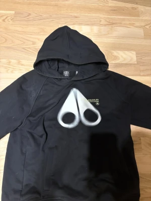 Svart hoodie från Moose Knuckles - Svart hoodie från Moose Knuckles med stor vit logga framtill och texten 'Engineered in Canada' i gult. Klassisk huva och känguruficka. Perfekt för dig som gillar streetwear och vill sticka ut med en unik design.