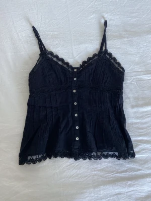 Svart spetslinnetopp från Zara - Snygg svart linnetopp från Zara med tunna axelband, spetsdetaljer längs kanterna och knappar framtill. Toppen har en feminin v-ringning och dekorativ spets både upptill och nertill. Justerbara axelband. Det finns ett litet märke på översta knappen (se bild) men inget man tänker på. Bortsett från det är den i toppen skick 💗