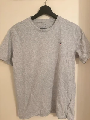 Ljusgrå t-shirt Gant - Säljer en stilren ljusgrå t-shirt från GANT med klassisk rund hals och liten logga. Den är storlek 176cm  i barn storlek alltså Xs-S.