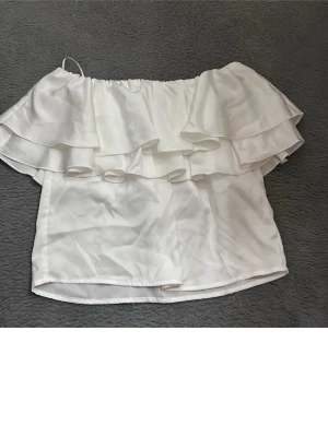 Vit offshoulder volangtop från Gina Tricot - Supersöt vit offshoulder topp från Gina Tricot med dubbla volanger. Perfekt för dig som gillar en luftig och trendig stil. Tillverkad i ett mjukt och lätt material som känns skönt mot huden. Passar perfekt till både jeans och kjol.