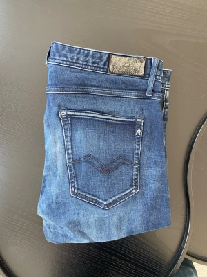Blå slim fit jeans från Replay - Säljer ett par blå slim fit jeans från Replay med snygga slitningar och klassisk femficksdesign. Jeansen har en mörk tvätt med ljusare partier och detaljerad bakficka med broderad logga. Perfekta för en trendig och avslappnad stil.
