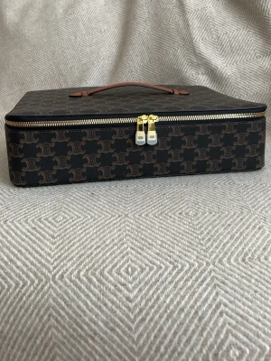 En brun sminkväska med monogram - Snygg sminkväska  i mörkbrunt med klassiskt Triomphe-monogram i ljusare brunt. Väskan har guldfärgad dragkedja och ett handtag i brunt läder. Rektangulär form och stilren design, perfekt för att bära din smink eller annat  med stil.