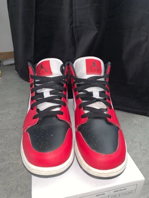 Air Jordan 1 Mid röd svart vit - Snygga Air Jordan 1 Mid sneakers i rött, svart och vitt läder. Klassisk siluett med svart swoosh, röda paneler och vita detaljer. Skorna har svarta skosnören och ikoniska Jordan-loggor på plös och sida. Perfekta för dig som gillar streetwear och basketstil.