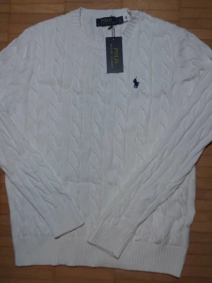 Vit kabelstickad tröja Polo Ralph Lauren - Vit kabelstickad tröja från Polo Ralph Lauren med rund halsringning och klassisk marinblå logga broderad på bröstet. Tröjan har ribbade muddar och är tillverkad i mjukt material som passar perfekt för en stilren look.