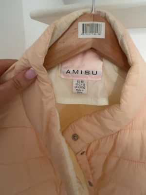 Ljusbeige dunväst från Amisu - Snygg ljusbeige dunväst från Amisu med quiltad design och tryckknappar framtill. Västen har en hög krage och är lätt vadderad, perfekt för lager på lager. Passar till många olika stilar och är superenkel att matcha. Den är i S
