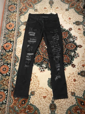 Svarta slitna jeans från Dsquared2 - Säljer ett par svarta jeans från Dsquared2 med slitningar och trasiga detaljer på framsidan. Jeansen har smal passform, klassisk femficksdesign och bälteshällor. Perfekta för dig som gillar en edgy och trendig stil. Storlek 44 = W28