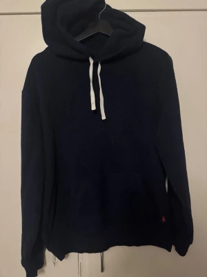 Mörkblå hoodie från Polo Ralph Lauren - Mörkblå Polo Ralph hoodie med huva . Klassisk känguruficka framtill och diskret röd logga broderad vid nederkanten. Perfekt för användning i vardagen och i vardagsrummet .