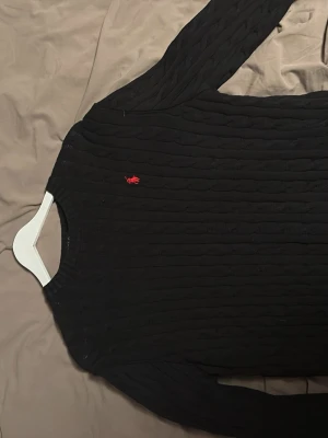 Svart kabelstickad tröja Polo Ralph Lauren - Kabelstickad svart tröja från Polo Ralph Lauren med rund halsringning och lång ärm. Klassisk röd logga broderad på bröstet. Tröjan har en slim fit passform och är perfekt för lager på lager. Materialet ser ut att vara mjukt och stretchigt.