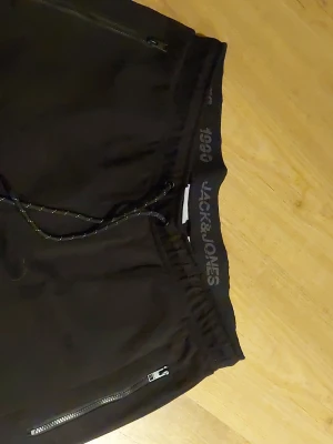 Svarta shorts från Jack & Jones - Svarta shorts från Jack & Jones med snörning i midjan och dragkedjeförsedda fickor. Elastisk midja med logga och sportig look. Perfekta för avslappnade dagar eller träning.