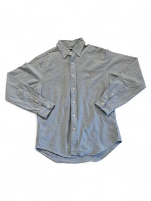 Grå skjorta från Ralph Lauren - Klassisk grå/blå skjorta från Ralph Lauren med broderad logga på bröstet. Skjortan har lång ärm, knappar framtill och krage. Tillverkad i mjuk bomull och passar perfekt till en avslappnad men ändå stilren look.