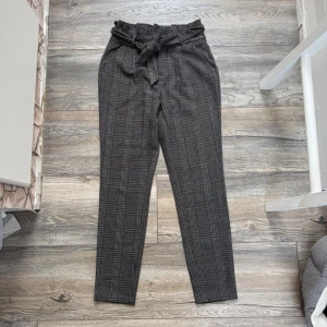 Rutiga kostymbyxor med knytskärp (Vero Moda, S) - Snygga kostymbyxor i grått med rutigt mönster och hög midja. Byxorna har ett brett knytskärp i midjan och raka ben. Perfekta för en trendig och stilren look. Materialet är mjukt och bekvämt. 
