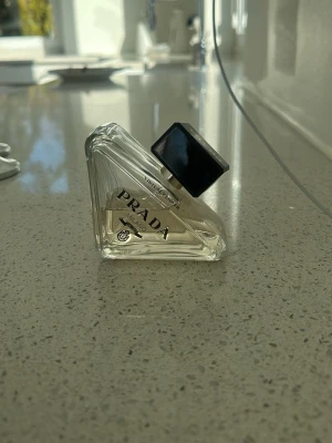Prada Parfym Triangle Flaska - Snygg parfym från Prada i en unik triangelformad glasflaska med svart fyrkantigt lock. Flaskan är transparent och etiketten har tydlig Prada logga. Perfekt för dig som gillar exklusiva dofter och stilren design.