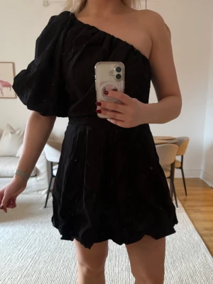 Svart oneshoulder klänning H&M - Svart kortklänning från H&M med oneshoulder-design och en puffärm. Klänningen har en markerad midja och är tillverkad i ett lätt tyg som ger en snygg siluett. Perfekt för dig som vill sticka ut med en trendig och stilren look.