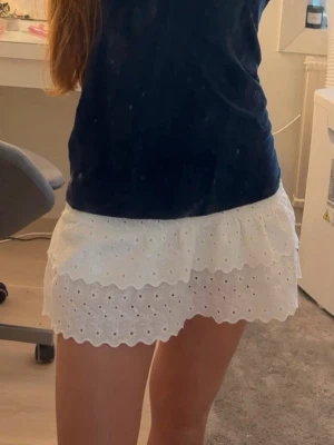 Vit volangkjol med broderade detaljer - Supersöt vit kortkjol med volang och broderade hål. Kjolen har två lager med vågig kant och ger en romantisk vibe. Perfekt till sommaren och lätt att matcha med olika toppar. Inbyggda shorts, pytteliten fläck högst upp på kjolen.