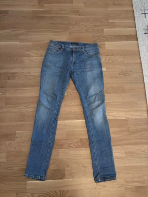 Blå slim fit jeans från Nudie - Säljer ett par klassiska blå slim fit jeans från Nudie med fem fickor och snygga slitningar. Jeansen har en normalhög midja och smal passform hela vägen ner. Materialet är mjuk denim i bomull med stretch för extra komfort.