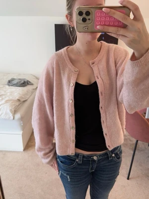 Ljusrosa kofta med pärlemorknappar - Mysig ljusrosa kofta med rund hals och pärlemorknappar. Koftan är stickad och har en lite lösare passform med långa ärmar och ribbade muddar. Perfekt att slänga över en topp för en chill look.