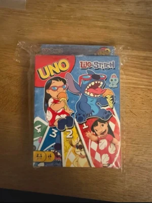 UNO Lilo & Stitch kortspel - UNO kortspel med Lilo & Stitch-tema. Färgglada spelkort med motiv av Lilo och Stitch som äter glass och leker. Förpackningen är blå, röd och gul med tecknade figurer från Disney. Perfekt för spelkvällar med vänner eller familj.
