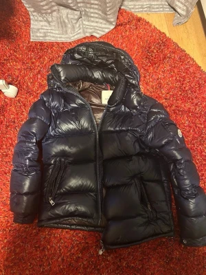 Mörkblå pufferjacka från Moncler - Snygg mörkblå pufferjacka från Moncler med glansig finish och stor huva. Jackan har två dragkedjeförsedda fickor framtill och Moncler-logga på ärmen. Perfekt för kalla dagar och riktigt trendig look.