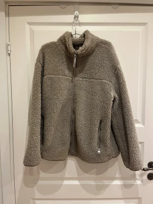 Beige teddyjacka med dragkedja - Mysig teddyjacka med hög krage och hel dragkedja framtill. Jackan har två fickor med dragkedja och är tillverkad i fluffigt teddy-material.