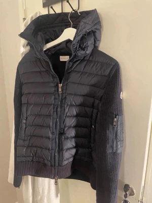 Svart pufferjacka från Moncler - Snygg svart pufferjacka från Moncler med stickade ärmar och huva. Jackan har dragkedja framtill, två fickor med knappar och en ärmficka med dragkedja. Perfekt för kalla dagar och riktigt trendig look.