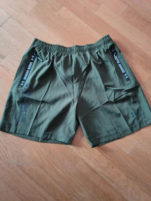 Gröna shorts från Under Armour - Snygga gröna shorts från Under Armour med elastisk midja och dragkedjeförsedda fickor. Shortsen har logga och textdetaljer på sidorna och är perfekta för träning eller chill. Tillverkade i ett lätt och bekvämt material.