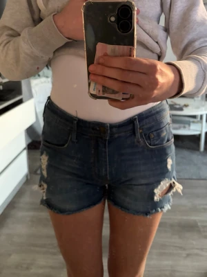 Mörkblå slitna jeansshorts - Snygga mörkblå jeansshorts med slitna detaljer och fransig kant. Klassisk femficksmodell med knapp och dragkedja framtill. Perfekta för varma dagar och en avslappnad stil.