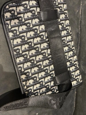 Svartvit axelväska/ midjeväska   - Säljer en snygg svartvit axelväska med klassiskt monogrammönster. Väskan har justerbar axelrem i textil och detaljer i svart skinn. Perfekt för dig som vill ha en stilren och ikonisk accessoar.