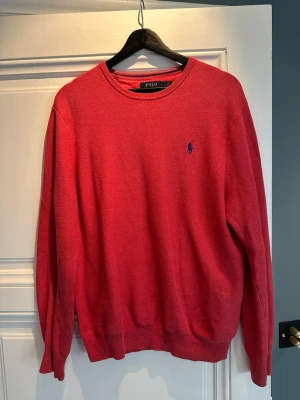 Röd tröja från Polo Ralph Lauren - Röd stickad tröja från Polo Ralph Lauren med rund halsringning och blå broderad logga på bröstet. Tröjan har långa ärmar och ribbade muddar vid ärmslut och nederkant. Perfekt för en stilren och klassisk look.