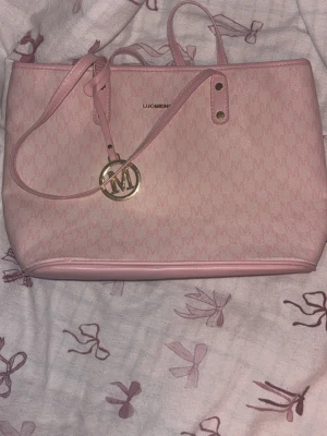 Ljusrosa hand/axelväska - Snygg ljusrosa hand/axelväska Från Shein med mönstrad yta och guldfärgad logga. Väskan har dubbla handtag, dragkedja på baksidan och dekorativt M-hänge. Tillverkad i konstläder med rosa detaljer och rymlig insida. Liten fläck inuti hade pennor i och korken hade lossnat så blev ett streck.