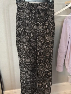 Svarta mönstrade byxor med vida ben - Säljer ett par svarta byxor med vitt grafiskt mönster och vida ben. Byxorna har hög midja och är tillverkade i ett mjukt, stretchigt material som ger en avslappnad look. Perfekta för dig som gillar statement-plagg och vill sticka ut.