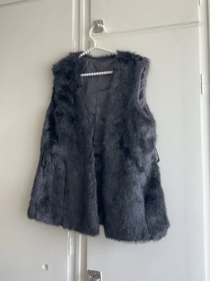 Mörkgrå pälsväst i fuskpäls - Snygg mörkgrå pälsväst i mjuk fuskpäls med slät insida. Västen är ärmlös och har en rak passform som ger en trendig look. Perfekt att slänga över en hoodie eller långärmad tröja för extra stilpoäng.