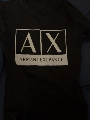 Svart t-shirt från Armani Exchange - Snygg svart t-shirt från Armani Exchange med stor vit logga framtill. Klassisk passform och rund halsringning, perfekt för en stilren och modern look. T-shirten är i mjuk bomull och har korta ärmar.